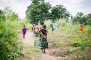 People walk along a rural path through cultivated fields while carrying young children.